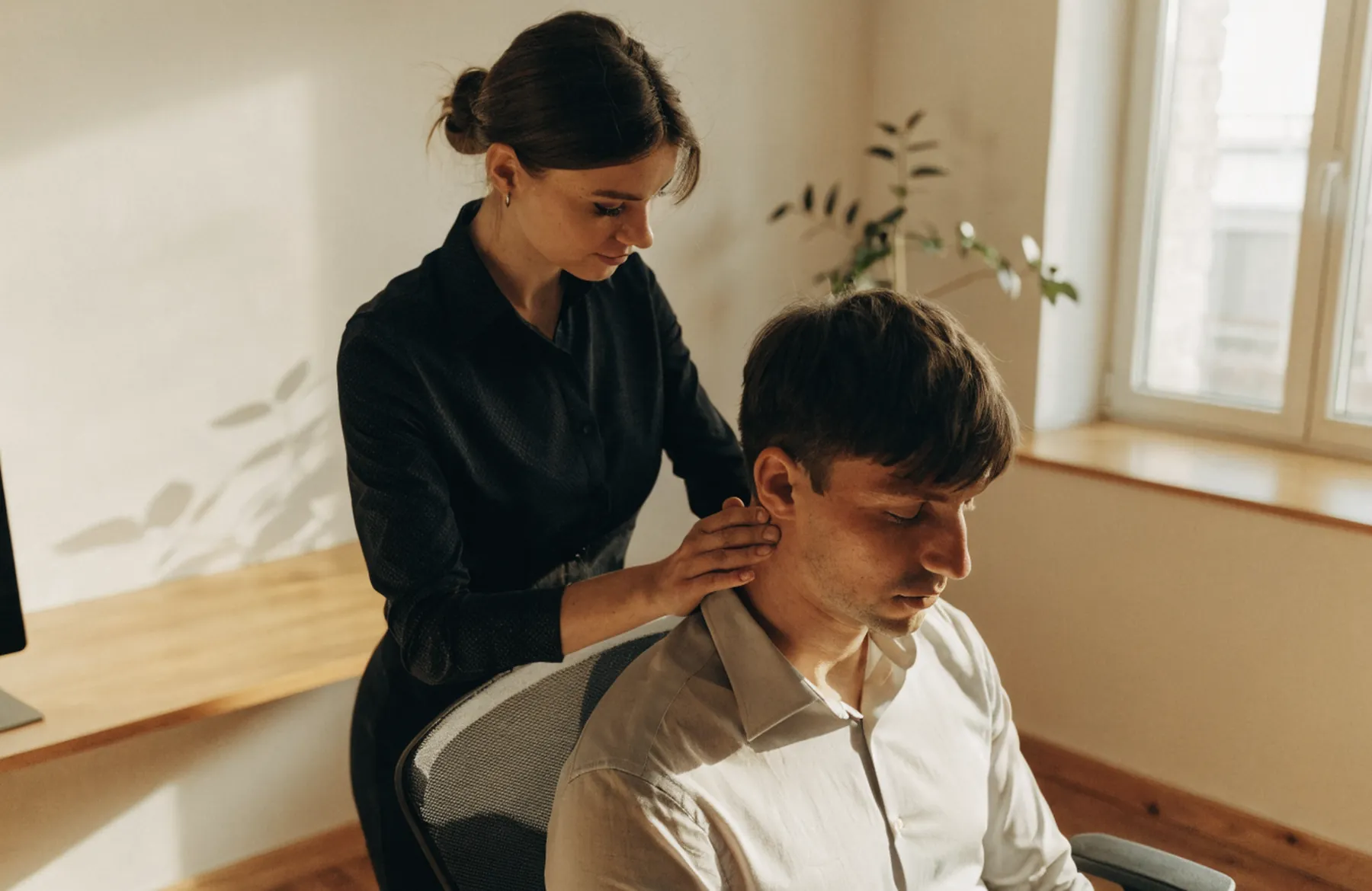 Terapeuta pracuje z górną częścią pleców siedzącego pracownika w jasnym pomieszczeniu